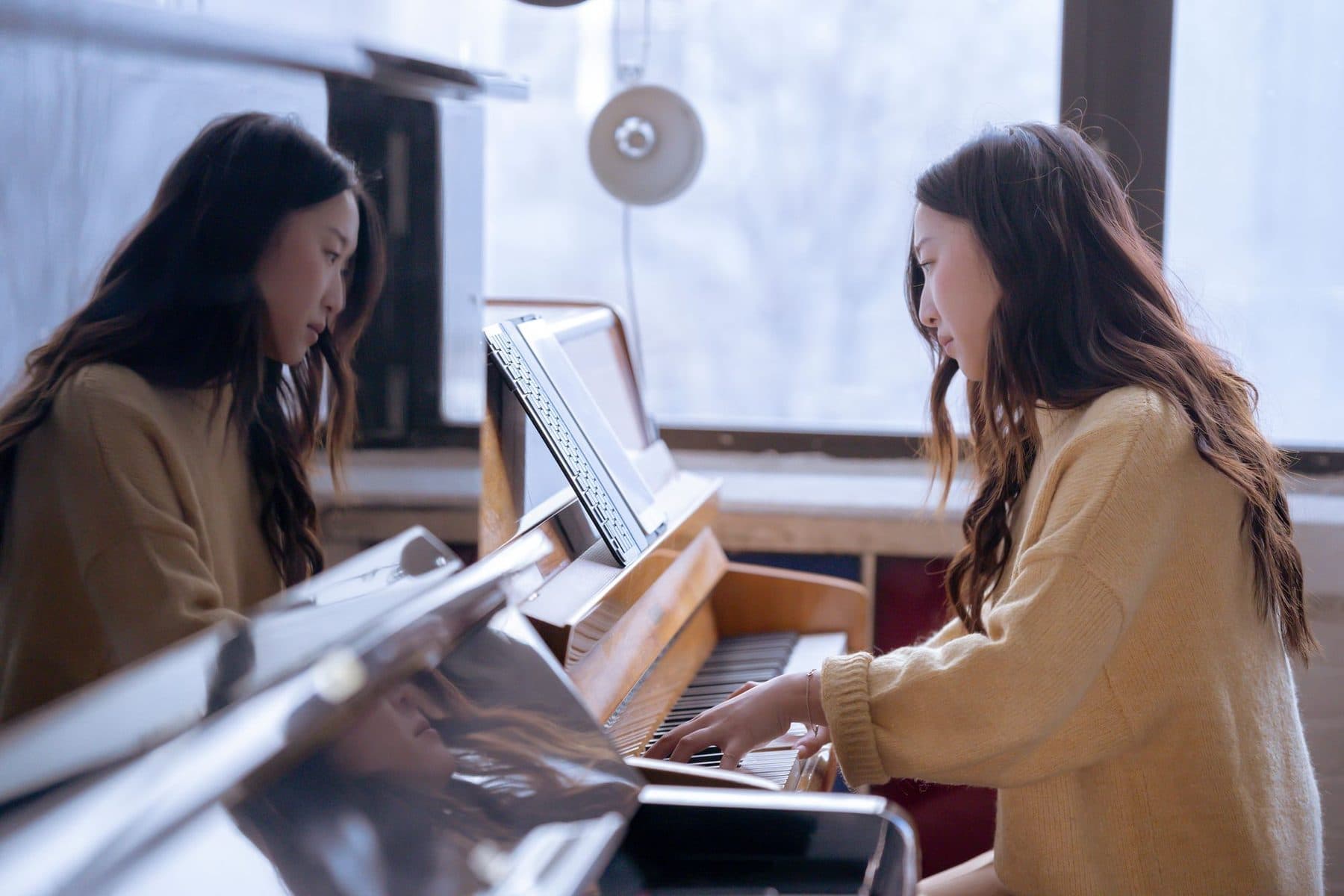 A young woman seated at a grand piano in a bright room, one hand on the keys.